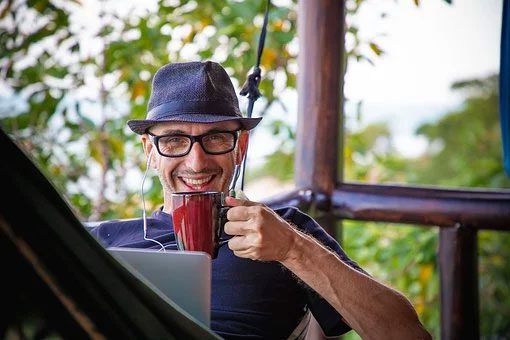 Man smiling holding cup wearing earplugs in hammock with laptop
